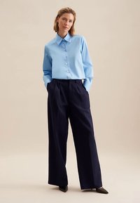 Camicia azzurra chiusa con bottoni, con colletto a punta, abbinata a pantaloni ampi color blu navy e scarpe nere con punta a punta. Tessuti con texture liscia.