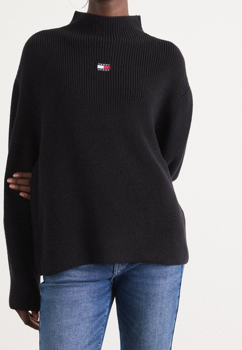 Personne portant un pull noir en maille côtelée avec un patch logo Tommy Jeans sur la poitrine, associé à un jean en denim bleu sur un fond uni.