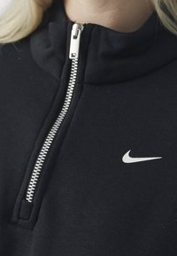 Černá zipová mikina vyrobená z měkké tkaniny, s stříbrným zipem a bílým logem Nike na hrudi. Vysoký límec.