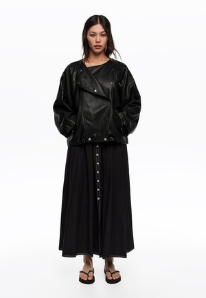 Kobieta o długich ciemnych włosach, ubrana w luźną czarną skórzaną kurtkę, długą czarną spódnicę zapinaną na guziki oraz czarne klapki, stojąca na białym tle.