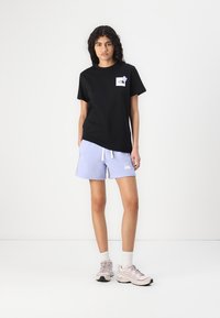 Črna majica s majhnim belim logotipom, v kombinaciji s svetlo vijoličnimi kratkimi hlačami, ki imajo bel emblem. Model nosi svetle športne copate z belimi nogavicami.