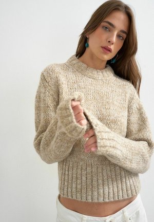 Jonge vrouw met lang haar, gekleed in een beige grofgebreide trui en witte broek, mouw aanscherpend, met turquoise druppelvormige oorbellen.