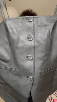 Haut sans manches en cuir gris à boutons avec deux poches devant, tenu par de fines bretelles sur un fond neutre.