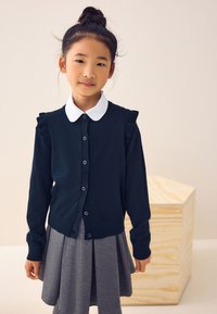 Granatowy kardigan z falbankami na ramionach, zapinany na guziki z białym kołnierzykiem, zestawiony z szarą plisowaną spódnicą. Gładka faktura, casualowy styl.