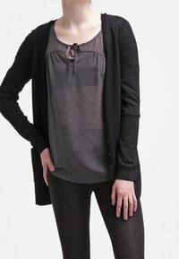 Svart cardigan över en grå blus med knytkrage, parat med svarta leggings. Tyget verkar mjukt, med en slät textur.