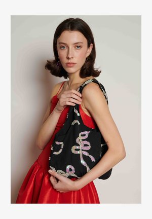 Giovane donna in abito rosso che tiene una borsa nera con motivi di paillettes lucenti, in piedi davanti a uno sfondo chiaro e semplice.