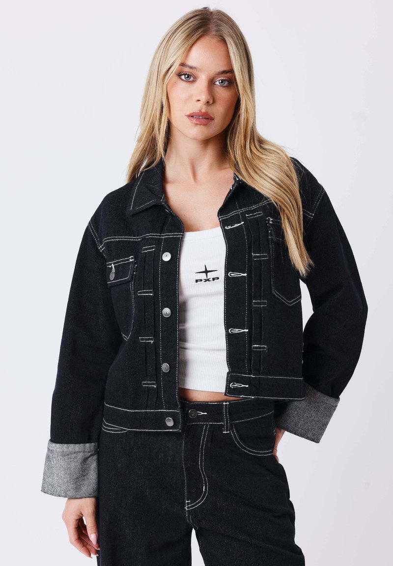 Femme blonde portant une veste en jean noire avec des poignets gris, un jean noir assorti et un top court blanc avec un petit logo noir.