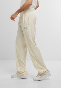 Pantalones de chándal color crema con una cintura elástica, bolsillos laterales y logo bordado. Tela suave con un ajuste relajado y diseño de pierna ajustada.