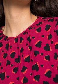 Fuchsiasnoppad topp med svart och orange hjärtmönster, med rundad halsringning och plisserade detaljer runt kragen. Slät tygtextur.