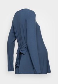 Pitkähihaisessa sinisessä paidassa on pehmeä kangas, jossa on solmukohta takana, pyöreä kaula-aukko ja rento istuvuus, joka levenee helmaa kohti.