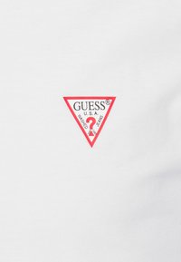 Hvid t-shirt med et trekantet rødt GUESS® logo. Logoet inkluderer teksten "U.S.A." og "WASHED JEANS", præsenteret centralt.
