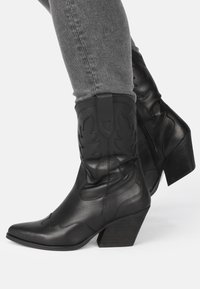 Botas de vaquero de cuero negro con punta afilada, costuras decorativas y un tacón ancho medio. Presentan un zipper lateral para facilitar su uso.