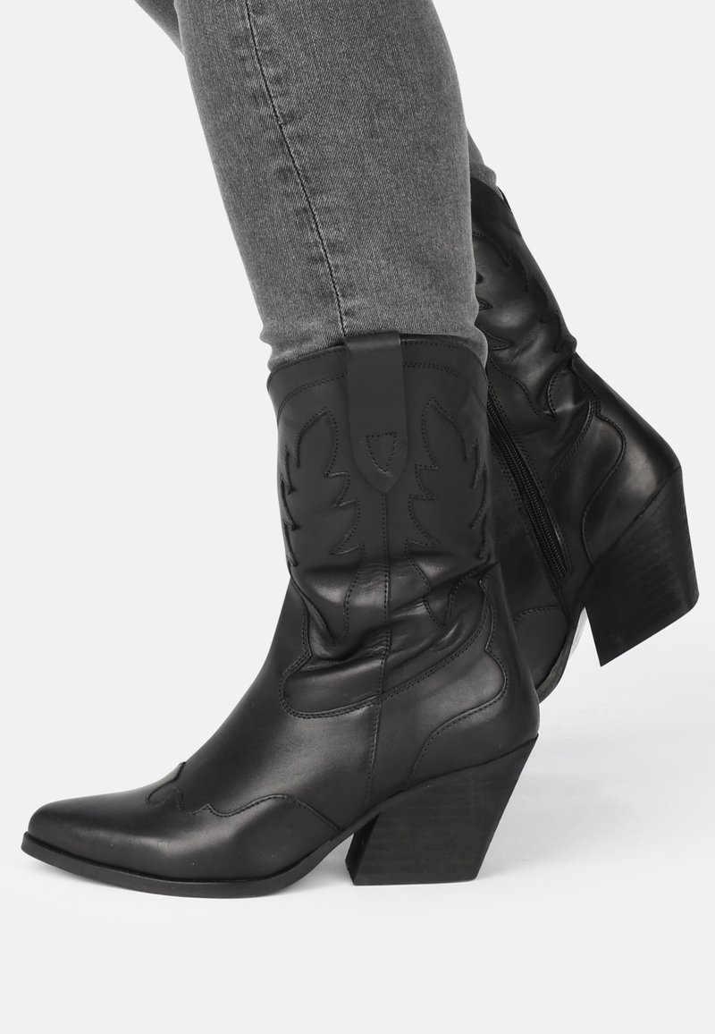 Botas de vaquero de cuero negro con punta afilada, costuras decorativas y un tacón ancho medio. Presentan un zipper lateral para facilitar su uso.