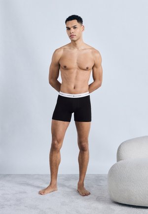 Ung mand stående barfodet iført sorte boksershorts med hvid Tommy Hilfiger taljebånd mod en ensartet baggrund.