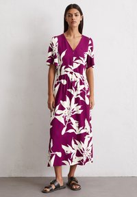 Lila Blumenkleid maxi mit kurzen Ärmeln, V-Ausschnitt, weißen Blättermustern, gepolsterter Taille und kombiniert mit schwarzen Sandalen.