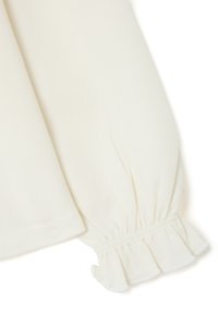 Long-sleeve top with a soft, cream fabric. Features a gathered sleeve cuff with subtle ruffle detail and a straight hem.