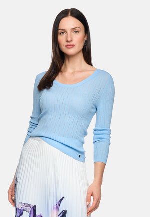 Woman with long brown hair wearing a light blue ribbed sweater and white pleated skirt with purple floral design, posing against a white background.