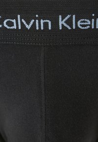 Schwarze Baumwoll-Stretch-Leggings mit einem strukturierten Bund, der mit einem hellblauen gestickten "Calvin Klein"-Logo verziert ist. Einfaches Design, nahtloser Abschluss.