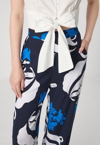 Blusa branca cropped com um laço nas costas, combinada com calças azul-marinho com grandes padrões florais brancos e azuis.