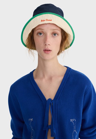 Jeune personne à la peau claire et cheveux roux portant un pull bleu avec une broderie d'oie et un chapeau bob beige et vert avec des lettres rouges.