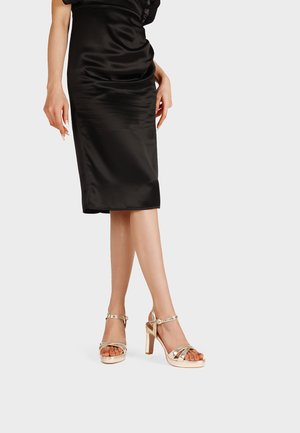 Donna che indossa un vestito di raso nero lungo fino al ginocchio e sandali dorati con tacco alto e cinturini alla caviglia, in piedi davanti a uno sfondo semplice.