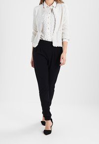 Biały teksturowany żakiet nałożony na koszulę na guziki w nadruk, zestawiony z ciemnymi spodniami i czarnymi szpilkami. Strój wyróżnia się subtelnymi wzorami i detalami.