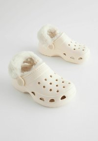 Sabots en crème clair avec doublure en fourrure, trous de ventilation et bords texturés. Forme ronde avec des boutons décoratifs sur le côté.