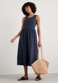 Donkerblauwe mouwloze jurk met een geraffineerde taille, rok tot op de knie, gecombineerd met een beige gestructureerde handtas en zwarte schuifsandalen.