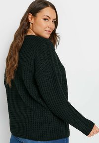 Svart stickad tröja med ett texturerat mönster, lös passform och ribbade långa ärmar. Baksidan är synlig och visar materialets tjocklek.