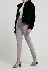 Zwarte fluwelen pufferjack met ritssluiting en drukknoopjes, grijze tapering broek met zijstrepen en zwart-witte gestreepte blokhak mules.