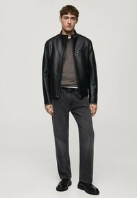 Giacca di pelle nera con colletto alto e zip frontale, t-shirt grigia, jeans grigio scuro e scarpe nere, in piedi su uno sfondo neutro.