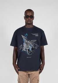 Marineblå bomulds t-shirt med et grafisk print af engleagtige figurer i blå og hvid, med "SAINT" trykt på forsiden.