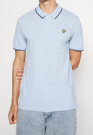 Mężczyzna w jasnoniebieskiej koszulce polo z czarną lamówką na kołnierzyku i rękawach, z haftowanym żółtym logo ptaka na piersi, w połączeniu z wyblakłymi niebieskimi dżinsami.