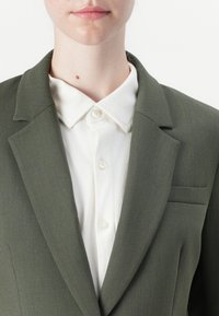 Olivgrön blazer med kammussla, en enda bröstficka och slät textur, lager på en vit knappad skjorta.