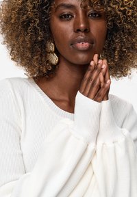 Vrouw met krullend haar en gouden oorbellen, draagt een witte geribbelde trui, handen samengevouwen dichtbij haar gezicht tegen een effen achtergrond.