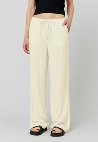 Pantalons larges, légers, couleur crème, avec une taille à cordon, dotés de poches latérales et d'une texture lisse. Portés avec des sandales noires.