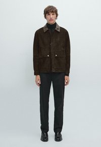 Giacca in suede marrone con chiusure a scatto, dotata di due tasche frontali e colletto a contrasto. Indossata sopra un dolcevita nero e pantaloni scuri.