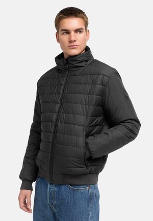 Junger Mann in einer schwarzen gesteppte Jacke mit Reißverschluss, eine Hand in der Tasche, seitlich schauend vor einem schlichten hellen Hintergrund.