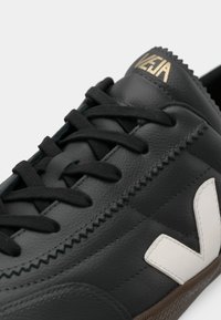 Sapatilha preta em pele com um logo V branco, detalhes texturizados, atacadores pretos e uma sola de borracha castanha. Os detalhes incluem costura em ziguezague.