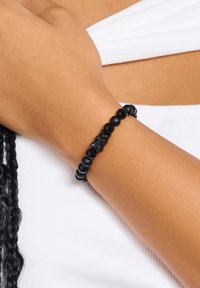 Black beaded bracelet featuring varied matte and glossy beads, including a central textured piece. Worn on a wrist, skin tone visible.