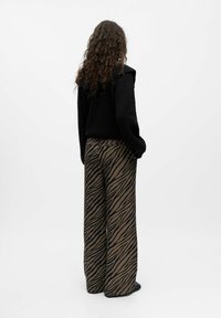 Personne avec de longs cheveux bouclés portant un pull noir et un pantalon marron à jambes larges ornés de rayures noires de tigre, debout les mains dans les poches.