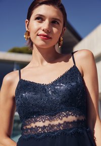 Robe de couleur marine avec des accents en dentelle et des sequins, présentant des bretelles spaghetti ajustables et une partie centrale transparente. De grandes boucles d'oreilles dorées complètent le look.
