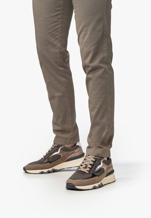 Nogi w brązowych dopasowanych spodniach i wzorzystych sneakersach w brązowo-czarnym odcieniu z beżem stojące na białej powierzchni.