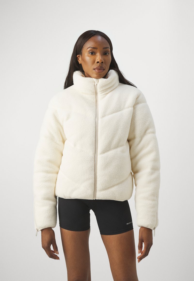 Weißer, flauschiger Fleece-Jacke mit hohem Kragen, Reißverschluss und strukturiertem geometrischen Muster; kombiniert mit schwarzen Shorts.
