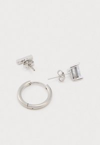 Ασημένιο σκουλαρίκι με λείο φινίρισμα δίπλα σε σκουλαρίκι με στύλο που διαθέτει ορθογώνιο πολύτιμο λίθο. Και τα δύο κομμάτια παρουσιάζουν καθαρούς, σύγχρονους σχεδιασμούς.