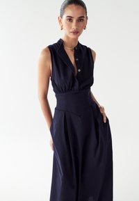 Temno modra brezrokavna kombinezona z gumbnim ovratnikom, prilegajočim pasom ter širokimi hlačami s ploščami in teksturirano tkanino.