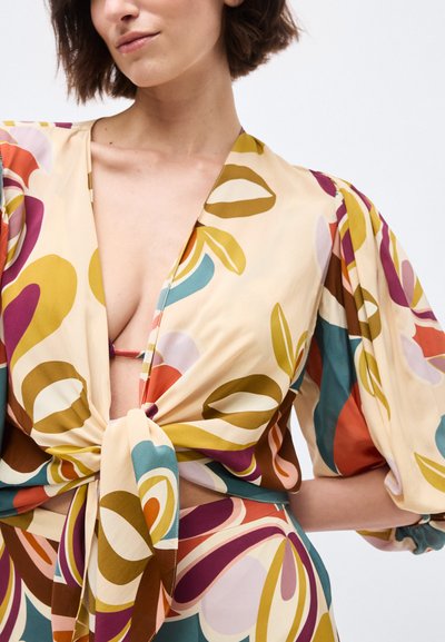 Γυναίκα που φορεί πολύχρωμη μπλούζα με αφηρημένο μοτίβο, φουσκωτά μανίκια και κόμπο μπροστά, με βαθύ V ντεκολτέ και φούστα με μοτίβο.