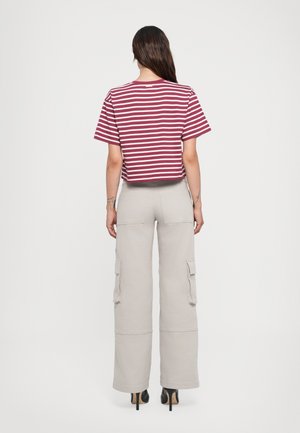 Femme debout de dos portant un crop top rayé rouge et blanc à manches courtes et un pantalon cargo beige clair à jambes larges avec des talons hauts.