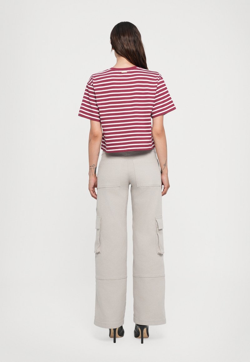 Femme debout de dos portant un crop top rayé rouge et blanc à manches courtes et un pantalon cargo beige clair à jambes larges avec des talons hauts.