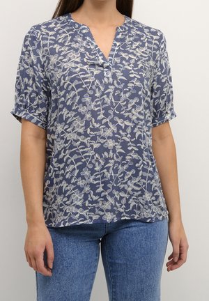 Kvinde iført en kortærmet blå bluse med hvide blomsterprint og blå jeans, stående mod en ensfarvet lys baggrund.